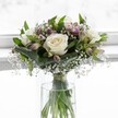 An elegant white rose bouquet rests in a clear glass vase, positioned in front of a softly glowing window that floods the scene with gentle daylight, like a quiet morning in a calm Tufnell Park townhouse. Three fully open, creamy white roses command the centre of the arrangement, their velvety petals curled outward in perfect spirals, revealing soft ivory hearts that suggest purity and new beginnings. Around them, layers of lush green foliage form a supportive cradle: broad, slightly folded leaves curve protectively, adding depth and a rich emerald frame that balances the lightness of the blooms. Threaded delicately between the roses and greenery are fine sprays of baby's breath, each tiny white flower hovering like a little cloud, giving the bouquet an airy, almost ethereal veil. Slender stems of alstroemeria emerge in muted shades of mauve and blush-pink; some buds are still closed, others just beginning to open, hinting at future colour and adding a sense of quiet anticipation. The stems are clearly visible through the cylindrical glass vase, immersed in clear water, neatly gathered and secured with a subtle ribbon just above the waterline, showcasing careful, professional floristry. The overall shape is compact and rounded, yet never crowded, with each bloom given space to breathe. The light, filtered as though through a white curtain on a side street off Fortess Road, lands gently on the petals and glass, highlighting delicate textures and the freshness of the flowers. This serene arrangement feels perfectly suited for a thoughtful condolence, a peaceful wedding detail, or a simple gesture of care, bringing a sense of calm, natural harmony into any Tufnell Park home.