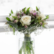 A calm, contemplative bouquet sits in a clear glass vase, its soft tones and gentle textures evoking a sense of quiet support, the kind often needed in times of loss around Tufnell Park and the nearby streets leading towards Archway. At the heart of the arrangement, several white roses are fully open, their velvety petals layered in generous spirals that move from creamy ivory at the centre to a delicate, luminous white at the edges. These blooms are wrapped in a light haze of gypsophila, the tiny white baby's breath flowers forming a fine, cloud-like mist that softens every line and edge. Threaded throughout are slender alstroemeria buds and blossoms, their elongated petals dusked with lavender and pale purple, some lightly striped, introducing a restrained touch of colour that never disturbs the bouquet's serene character. Around the base of the flowers, broad, glossy dark-green leaves have been carefully folded and wrapped, creating a smooth, sculpted cradle that holds the rounded, hand-tied form together. Additional narrow green leaves rise between the blooms, adding movement and a sense of gentle life. Inside the cylindrical vase, clear water surrounds the long green stems, which are drawn together neatly with a small band of green ribbon or raffia just above the waterline, ensuring the bouquet maintains its graceful, domed shape. The softly lit, pale background fades into a bright blur, as if the flowers are placed near a sunlit window in a quiet North London home. The arrangement's balanced composition, soft whites, and cooling greens convey respect, comfort, and enduring love, making it an especially thoughtful choice for sympathy deliveries within the local area.