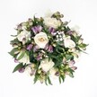 Top-down view of a hand-tied bouquet laid against a crisp white background, its rounded shape filled with creamy white roses, deep purple alstroemeria buds, clusters of white baby's breath, and glossy dark green foliage, arranged with the quiet elegance of a Tufnell Park florist. At the centre, several full, velvety roses in shades of ivory and pale champagne open in soft spirals, while a few remain partially unfurled, their petals cupping inward as if mid-breath. Threaded between them, alstroemeria buds in rich violet, plum, and magenta emerge from fresh green bases, their tapered forms adding rhythm and movement to the design. Tiny clouds of gypsophila flicker throughout like a delicate floral mist, lightening the deeper tones and giving the bouquet a gentle, celebratory air. Around the outer edge, broad, polished leaves create a lush frame, their dark emerald surfaces catching the soft, even lighting. The absence of shadows allows every petal, vein, and curve to be seen clearly, conveying that just-arrived freshness you might notice when flowers are delivered to a townhouse off Fortess Road or a flat overlooking Tufnell Park playing fields. This bouquet feels balanced yet organic, refined yet warm, making it easy to imagine it cradled in someone's hands on an anniversary, a quiet celebration at home, or a thoughtful get-well visit. The image holds a mood of calm sophistication and gentle romance, a still moment where colour, texture, and tenderness meet.