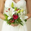 In this close-up bridal scene, a hand-tied bouquet rests against a lace wedding dress, creating a tender contrast between delicate fabric and living flowers that could easily be imagined in a Tufnell Park church or town hall ceremony. The composition is built around a single, perfectly formed red rose, its deep crimson petals spiralling inward with velvety richness, symbolising enduring love. Surrounding this focal point, large white lilies open in graceful starbursts, their creamy petals curling slightly at the edges and catching the light with a soft sheen. Some of the lilies are at full bloom, their pollen-laden stamens visible, while others remain in smooth, elongated buds, lending an air of unfolding promise. Interwoven among them are airy sprigs of gypsophila, each tiny white blossom light as a sigh, echoing the intricacy of the lace gown. Clusters of bright red berries punctuate the design, shiny and festive, balanced by tufts of fresh green moss and dianthus that introduce a gentle woodland feel reminiscent of the greenery around Tufnell Park's quiet residential streets. Fine blades of ornamental grass and feathery foliage extend outward, giving the bouquet a soft halo effect and a sense of movement, as though caught in a moment of happy anticipation. Held in the bride's hands, the stems are neatly gathered and wrapped in a subtle ribbon, almost hidden from view. The bouquet radiates romance and refined joy, a thoughtful blend of purity, passion, and texture that would feel completely at home in an intimate North London wedding, lovingly prepared for same-day bridal flowers in and around Tufnell Park.
