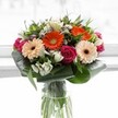 Imagine stepping into a sunlit Tufnell Park kitchen, where this vibrant bouquet instantly commands attention with its interplay of colour and form. At the heart, gerbera daisies in rich orange and creamy peach offer a sense of warmth and cheer, each bloom's brown centre striking a gentle contrast. Surrounding them, deeply magenta roses evoke both passion and sophistication, their velvety petals appearing impossibly fresh. Sprigs of white alstroemeria and tiny filler flowers peek from behind, scattering light and softening the overall look, much like the first daisies of spring along Parkhurst Road. Cradling the blooms, aspidistra leaves are wrapped with care to create a supportive, verdant collar, while slender, pointed foliage punctuates the bouquet with lively green notes. The clear vase below reveals a tapestry of vibrant stems, amplifying the bouquet's sense of organic vitality. Sunlight pours in from behind, illuminating the arrangement and inviting thoughts of new beginnings, local celebrations, or simply brightening a day for someone cherished. This lavish creation, shaped by local expertise, brings a taste of Tufnell Park's community spirit-ideal for gifting at home, in a favourite café, or to send a message of affection across North London.