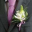 A close-up, high-angle photograph shows the upper torso of a person dressed for a formal celebration in Tufnell Park, the dark charcoal grey of their tailored suit jacket creating a refined backdrop for a single, beautifully crafted buttonhole. The lapel is sharply cut and pressed, framing a pale dress shirt with a fine grid pattern that peeks from beneath the open jacket. Down the centre runs a smooth lavender tie, its sheen catching soft indoor light much like the evening glow you might see along Brecknock Road before a wedding reception. Pinned carefully to the left lapel, the boutonniere takes centre stage: a slender, pale green spire of unopened buds rises at the top, giving vertical elegance and a sense of promise. Around it cluster white alstroemeria blooms, their petals creamy and fresh, each one brushed with delicate streaks of maroon and hints of golden stamens at the throat. Tiny clouds of white gypsophila nestle between the larger flowers, lending a misty, romantic softness that feels perfect for a ceremony in a local church or registry office near Tufnell Park station. Slender, grass-like blades arc gently around the flowers, interwoven with broader, glossy green leaves that add depth and contrast against the dark fabric. At the base, a neat bow in satin lavender ribbon mirrors the tie exactly, its folded loops and cut ends holding the stems in a graceful, polished finish. The overall impression is one of quiet confidence and fresh anticipation, capturing that still, intimate moment before stepping out to meet waiting guests, with every detail of the florals carefully composed as though by an experienced local florist who knows how to balance elegance and emotion.