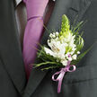 In this detailed close-up, the viewer is invited to linger over the harmony between formal wear and floral design, as if standing in a sunlit doorway of a Tufnell Park townhouse just before guests head to the ceremony. The frame is cropped to the torso, showing only the charcoal grey suit jacket, the shirt, the tie, and the carefully pinned boutonniere. The jacket itself is impeccably tailored, its lapel pressed flat with a faint sheen that speaks of quality cloth and good preparation. Beneath it, a light dress shirt with a fine dark check pattern adds an almost woven texture, subtle but stylish. A solid lavender tie in smooth satin sits proudly at the centre, its knot symmetrical and precise, reflecting a gentle, cool-toned light. The boutonniere is the star: several white alstroemeria blossoms open wide, their petals marked with russet speckles and warm yellow flashes at the throat, each flower finely veined. From the heart of the arrangement rises a single conical green spike, likely a tuberose or similar bud, its fresh lime-green tone offering height and a sense of newness. Around these, airy clusters of white gypsophila float like tiny stars, while a mix of slender grass-like blades, soft fronds, and deeper green leaves give body and contrast. A narrow lilac ribbon is tied neatly at the stem, echoing the tie's colour so precisely that the flowers feel made just for this outfit. The image captures that quiet, hopeful pause before a wedding, prom, or family celebration somewhere between Dartmouth Park and Kentish Town, with each floral choice reflecting professional care and local florist know-how.