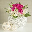 On a bright, minimal backdrop that feels like a sunlit apartment near Tufnell Park station, a graceful floral arrangement in a round white wicker basket creates a sense of quiet romance. At the bouquet's heart, vivid fuchsia blooms cluster together in rich, petal-heavy domes, their colour intense and velvety, reminiscent of lush peonies or garden roses at the peak of summer. These deeper pink flowers are embraced by a ring of ivory alstroemeria, each bloom with fine, painterly markings that add depth without overwhelming the eye. The petals curve outward gently, lending softness and a feeling of airiness, while glossy green foliage and a few emerging buds rise delicately through the composition, suggesting freshness and new beginnings. The wicker container is pure, brilliant white, its intricate latticework handwoven into a perfectly rounded shape that feels both contemporary and a little rustic-very much at home in the townhouses stretching toward Dartmouth Park. Two small, cream-coloured woven spheres rest beside the basket, repeating the basket's texture in a subtle, sculptural way, like thoughtful décor on a stylish wedding table in a local Tufnell Park venue. The lighting is diffused and gentle, cloaking the scene in a soft glow that picks up every curve of petal and strand of wicker. The overall feeling is of poised celebration: modern, elegant, and quietly joyful, ideal for a wedding collection piece, a bridal shower, or an engagement dinner. You can almost imagine the crisp, green fragrance and the soft whisper of leaves, captured with the kind of care a local florist brings to each handcrafted wedding design.
