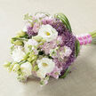 A meticulously crafted bouquet is shown resting on a soft, light beige cloth with a gentle woven texture, giving the arrangement the feel of being placed on a linen-dressed table in a calm Tufnell Park home. The flowers themselves form a horizontal spray, dominated by full, creamy white lisianthus whose layered petals open in generous circles, revealing subtle yellow-green centres that glow gently against the pale backdrop. Some lisianthus buds remain partially closed, their tight swirls adding structure and a sense of unfolding moments. Nestled among them are clusters of soft pink and lilac stock flowers, each dense spike made of dozens of tiny, ruffled blooms that lend a romantic, almost cloudlike volume and a faintly sweet, nostalgic scent. Punctuating the pastels are striking clusters of star-shaped purple flowers, similar to ornamental allium, with their fine, spiky heads creating a lively contrast in both colour and texture, like small bursts of lavender firework against the white and blush tones. Long, narrow blades of deep green foliage curl and loop gracefully through the design, especially along the right side, adding movement and drawing the eye along the bouquet's length, much like the gentle sweep of greenery you might see strolling towards Tufnell Park station or along Fortess Road. The stems are pulled together in a tight, elegant bind wrapped with vibrant fuchsia satin ribbon, the bold colour echoing the pink blooms, and overlaid with a delicate strand of tiny white pearls. This careful finishing detail makes the bouquet ideal for a wedding, anniversary, or special delivery around Tufnell Park, exuding understated luxury and heartfelt celebration.