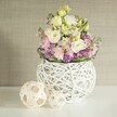 A celebration of Tufnell Park's gentle elegance unfolds in this handcrafted floral centrepiece, designed with the quiet sophistication local residents cherish. At the heart is a round white vase, beautifully woven from delicate painted rattan, with its airy, intricate openwork adding modern lightness to the display. Overflowing from the vessel, ruffled lisianthus-creamy white and subtle yellow-spill outward, interspersed with slender stems of soft pink stock that reach skyward in a dance of colour and form. At the base, a cloud of rich purple filler-most likely petite asters-cushions the main blooms, while a circlet of deep green foliage frames the composition, echoing the verdant canopy of local parks in spring. The vase sits atop a flawless, luminous white surface that seems to glow with Tufnell Park's morning light, its warmth enveloping every detail. To the left, two decorative spheres crafted from pale, open-woven rattan add a welcoming touch, their organic forms paying homage to local artisanal traditions. The backdrop is a refined neutral beige, perhaps a softly textured wall covering, which lets the arrangement's pastel tones and crisp lines sing. Together, these elements create an ambiance of tranquility and handmade beauty, perfect for inspiring wedding décor at The Grafton or for bringing a breath of fresh air into family gatherings throughout Tufnell Park.