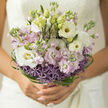 A hand-crafted bridal bouquet is held close at waist height, its pastel tones glowing gently against what feels like soft natural daylight drifting in from a north London window. The arrangement is composed of white lisianthus and pale blush stocks, their petals layered like fine silk and gathered together to form a rounded, slightly loose dome. Interlaced among them are touches of lilac, lending a cool, romantic hue that echoes the calm charm of Tufnell Park's tree-lined streets. At the base, delicate clusters of lavender allium create a ring of star-shaped florets, each tiny blossom contributing a subtle burst of purple and an almost celestial texture. Slim, glossy strands of bear grass sweep and curl around the bouquet's edge, looping into elegant arcs that give the design a contemporary, almost architectural outline while still feeling soft and romantic. The flowers look freshly cut, as if they've come straight from an early-morning delivery to a studio near Tufnell Park underground station, stems still cool to the touch. Gentle light catches the ruffles of each lisianthus petal, revealing hints of cream and soft ivory within the folds, while the stock blooms suggest a faint sweetness in the air-clean, floral, and uplifting. This bouquet feels ideal for an intimate wedding ceremony at a local registry office or a small gathering in a townhouse near Fortess Road, understated yet deeply expressive. Every spiral of grass, every carefully placed allium head speaks of a florist's steady, experienced hands, turning tranquil colours and simple shapes into a refined, quietly joyful composition that celebrates love without overwhelming the senses.