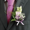 A close-up photograph focuses on the upper torso of a man in a sharply tailored charcoal suit, most likely a groom or honoured guest at a Tufnell Park wedding, standing in soft natural daylight. The jacket's textured lapel and sleeve sit smoothly over a pale dress shirt with a fine check pattern, while a vivid magenta silk tie runs down the centre, its sheen catching the light with a subtle gleam. Pinned neatly to the left lapel is a beautifully composed buttonhole, its colours and shapes carefully balanced by an experienced florist. At the top, slender buds of creamy white flowers, reminiscent of snapdragons just about to open, rise gracefully, giving height and poise. Nestled beneath them, small blooms in gentle pink and lavender-lilac tones - like sweet peas or delicate stock - cluster together in a rounded posy, their petals soft and almost velvety to the eye. Tiny clouds of white gypsophila weave through the arrangement, adding an airy, mist-like texture, while tucked hints of deeper purple florets create depth and quiet drama. Broad, glossy dark green leaves cradle the flowers, providing structure and contrast, supported by light, feathery sprigs of brighter green foliage that arc outwards in a relaxed curve, suggesting movement. At the base, a slim violet or lilac satin ribbon is tied in a tidy bow, its satin surface reflecting a soft glint and echoing the cooler floral tones. The overall impression is one of refined elegance and joyful anticipation, perfectly suited to a spring or summer ceremony in a local venue near Tufnell Park Road or the leafy paths of nearby Hampstead Heath, capturing the sense of new beginnings and celebration in a single, thoughtful flourish.