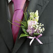 A single, artfully composed boutonnière rests against the rich, dark lapel of a tailored suit, its delicate colours bringing softness and romance to the formal attire. At its heart, pale lavender blossoms open like tiny cups of colour, their petals velvety and finely edged, surrounded by smaller creamy buds that lend a sense of gentle promise. Around them, airy tufts of white baby's breath form a halo of miniature blooms, as though a light mist has settled over the flowers. Slender strands of fresh greenery and fine grass lift the design upwards, giving the arrangement an elegant vertical line, while polished, curved green leaves cradle the composition below, offering a deep, natural contrast to the pastel flowers. A sheer lavender ribbon wraps the stems with quiet precision, its soft, satiny gleam picking up the tones of the nearby tie and subtly tying the whole outfit together. The scene feels intimate and calm, like a moment paused in the foyer of a townhouse near Tufnell Park station, just before the ceremony begins. You can almost sense the faint, sweet fragrance of the blooms, mixed with the crispness of freshly pressed fabric and the low hum of guests arriving. Crafted with the practiced hands of a local florist who understands both style and sentiment, this boutonnière becomes more than a simple floral accent: it is a small emblem of affection, celebration, and thoughtful preparation, perfect for weddings, proms, or special occasions across Tufnell Park and the surrounding North London neighbourhoods.