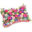 Seen from a gentle overhead, oblique angle against a clean white backdrop, this pillow-shaped floral tribute reads like a soft, wearable garden. The rectangular cushion is densely packed so no oasis of foam shows through: hot pink roses form the principal melody, their velvety petals in varied stages from tight buds to softly unfurling blooms. Between them, pale shell-pink carnations ripple with frilled edges, giving the surface a plush, tactile quality. Bright lemon-yellow ranunculus and cheerful buttercup-like sprays punctuate the composition, lifting the warmth of the pinks, while deep purple lisianthus adds pockets of cool contrast that draw the eye inward. Architectural accents of silver-blue eryngium thistles introduce a spiky, windswept note that plays against the softer textures, and small pink filler flowers scatter tiny, delicate details across the top. Rounding, silvery eucalyptus leaves and a mix of darker green foliage nestle throughout, providing a muted, verdant framework that lets the colours sing. A deep red, pleated satin ribbon frames the edge in a ruffled border, defining the cushion's silhouette and lending an understated elegance that suggests ceremonial use. The lighting is even and soft-like gentle morning light in a chapel or family home-so each petal's surface and shadow is visible without harsh glare. This arrangement is crafted with the quiet intent of remembrance: a vibrant yet tender sympathy pillow that celebrates a life with colour and texture rather than only sombre tones. It would be fitting for a memorial service or graveside tribute in Tufnell Park or nearby Dartmouth Park, where friends and family gather to remember with warmth.