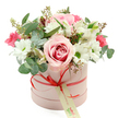 A softly glowing floral hatbox arrangement sits alone against a pristine white background, inviting the eye to linger on every detail. The container is a cylindrical hatbox in a gentle pastel pink, its surface matte and smooth, echoing contemporary gift packaging seen in stylish Tufnell Park boutiques. Around its middle runs a slim coral-red satin ribbon, tied into a neat, symmetrical bow at the front, from which a small cream tag hangs delicately. The tag features green script-like text and a modest web address, suggesting a quiet, local florist's mark rather than bold branding. Above the rim of the hatbox, the flowers rise in a rich, rounded dome that feels both lush and carefully composed. A striking pale pink rose, tinged with a deeper pink swirl at its heart, forms the central focal bloom, its petals layered and fully open, like a gently unfurling spiral. Clusters of clean white daisy chrysanthemums with vivid green centres surround it, introducing a fresh, joyful energy. Threaded between these are slender white alstroemeria, their trumpeted petals showing faint inner speckles that reward a closer look, while deeper fuchsia and rich pink flowers-probably spray roses or lisianthus-add vibrant flashes of colour that stop the arrangement from feeling too delicate. Silver-green eucalyptus leaves, round and softly textured, weave through the bouquet with darker green foliage and airy stems tipped with tiny buds, adding volume and movement. The composition feels ideal for a romantic surprise or an uplifting gesture, the kind of gracefully arranged hatbox someone in Tufnell Park might send to a friend near Campdale Road, bringing a sense of calm celebration and thoughtful elegance into their home.
