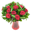 A close, intimate portrait of a classic romantic bouquet: roughly a dozen to fifteen deep crimson roses, each bloom full and velvety, their petals spiralling from tight centres into luxuriant, satin textures. The roses sit in a lush collar of dark green foliage-glossy, broad leaves like Salal or Ruscus form a dense backdrop while larger, emerald-green blades have been skillfully rolled and looped into architectural curls that cradle and lift the rose heads. The leafy loops are polished and reflective, their smooth surfaces catching soft, even studio light that emphasizes the contrast between the satin petal and glossy leaf. The arrangement is presented in a tall, translucent ruby-red glass vase with a subtle ribbed or twisted surface; vertical lines within the glass catch highlights and create a sense of upward movement that complements the rounded bouquet. Set against a clean white background, the saturated reds read almost wine-dark in places, and the greens feel deep and verdant, isolating the composition so every velvety swirl and folded leaf is visible. There's an implied scent of classic rose perfume-soft, warm, and lingering-while the bouquet's balanced volume and formal silhouette suggest a carefully considered gift for an anniversary or Valentine's moment. Imagine receiving this on a quiet morning in Tufnell Park, perhaps after a stroll past Fortess Road shops or a picnic near Dartmouth Park; the arrangement reads as both timeless and modern, a refined gesture that combines traditional romance with contemporary leaf styling and glassware that anchors it as a handsome centrepiece.