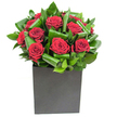 Rich, velvety red roses form the magnetising centre of this contemporary arrangement, each bloom opening in generous spirals that read like small, hand-forged roses of fabric and scent. The roses are tucked into a chorus of broad, glossy green leaves that have been artfully curled and interlaced; those emerald coils add a sculptural rhythm and a tactile contrast to the soft, satiny petals. The composition sits low and assured in a matte black, square box vase whose clean geometry offers a grounded, modern counterpoint to the organic movement above. Bright, even studio lighting catches the sheen on each leaf and the velvet highlights of the crimson petals, giving the bouquet a luminous clarity-as if it had been placed on a Tufnell Park café table during a crisp morning. Close to the nose, the scent is classic rose softened by green, almost citrusy notes from the foliage, a fragrance that breathes fresh and reassuring rather than sweetly overwhelming. As a florist describing this piece to someone who cannot see it, I would emphasise how the leaves frame and lift the roses, creating depth and a gentle heartbeat of motion across the arrangement. The overall mood feels intimate and deliberate, suited to an anniversary, a heartfelt thank-you, or to bring an elegant, romantic focus into a small urban living room or reception desk near Junction Road. The design's confident simplicity makes it a memorable gesture-modern, tactile, and quietly expressive without needing adornment.