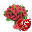 A richly detailed romantic gift set is displayed as if ready for hand-delivery to a doorstep in Tufnell Park, featuring a striking bouquet of red roses and a bold heart-shaped balloon. At the centre of the composition, a clear, fluted ruby-red glass vase holds a dense cluster of approximately one dozen or more deep crimson roses. The vase itself has subtle vertical ridges that catch the light, creating bands of darker and lighter red along its surface. Emerging from the neck of the vase, the roses appear in various stages of bloom: some blossoms are fully opened, showcasing plush, velvety petals layered in generous spirals, while others are just beginning to unfurl, their centres still tight and promising. The petals are a luxurious, uniform crimson, with soft curves that invite the imagination of fragrance and touch. Surrounding the blooms is a halo of rich green foliage, with leaves that are smooth and slightly glossy, offering a fresh contrast that anchors the bouquet in nature. In the lower right foreground, slightly overlapping the vase and drawing the eye, a shiny red heart-shaped foil balloon adds a playful, celebratory note. The balloon's metallic surface gleams, and across its centre, the message I Love You is written in graceful white cursive script, clearly visible and emotionally direct. A silver Cupid's arrow graphic appears to pierce the balloon diagonally, underscoring the theme of love and devotion. Set against a simple, solid white background, the vivid reds and greens stand out dramatically, making this image ideal for representing Valentine's bouquets, anniversary flowers, or romantic rose deliveries across Tufnell Park and nearby streets.
