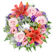 A generous, circular funeral wreath is shown from directly above, resting visually on a clean white background so that every flower and leaf is clearly defined. The design is lush and balanced, with no empty spaces, giving a sense of abundance and care. Throughout the ring, clusters of large, velvety roses in blush and light lavender-pink unfold in full bloom, their soft petals layered in delicate spirals that suggest comfort and affection. Nestled among them are lisianthus (eustoma) in gentle pinks and deep purples, their ruffled blossoms introducing a flowing, almost painterly texture. Two striking orange-red Asiatic lilies, one near the top and one near the bottom of the circle, serve as visual anchors, their wide, star-shaped petals and prominent stamens adding a warm glow to the composition. Threads of airy, lavender-blue filler flowers, probably statice, drift through the arrangement like a cool mist, linking the different shades together. The base foliage is rich and varied: rounded silvery eucalyptus, dark green fern fronds, and glossy leaves provide contrasting shapes, while blue-grey eryngium thistles, with their spiky, sculptural heads, give the wreath a subtle contemporary edge. Several unopened lily buds rest quietly within the greenery, promising flowers still to come, like cherished memories that linger. Bright green, striped or folded leaves, perhaps Ti or aspidistra, are carefully placed around the lilies, framing them with a modern touch. The overall impression is one of calm reverence and gentle beauty, a wreath you might see delivered to a Tufnell Park chapel or graveside, offering a respectful, heartfelt expression of sympathy and enduring love.