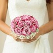 There's a gentle joy radiating from this scene set along the tranquil byways of Tufnell Park-a bride, poised and serene, holds a round bouquet that glows with nuanced pinks. At its heart: a dozen roses with petals curling inwards from a creamy blush to a vivid fuchsia, each bloom perfectly shaped and responsive to the morning's cool light. Wispy clouds of baby's breath, gathered from nearby markets, thread between the roses, their tiny, white blossoms adding a soft veil-like effect that conjures sentiments of innocence and hope. The bouquet sits nestled against a sleeveless white lace gown, the intricate floral patterns and a loosely tied sash echoing the old-world charm of Tufnell Park's Victorian terraces. The bride's hands, golden in tone, gently cradle the arrangement, her engagement ring and shimmery nude manicure catching the light. In the background, a softened canvas in warm beige ensures every detail of the bouquet sings. Each petal, every sprig of gypsophila, is evidence of a florist's care and artistry-crafted for a once-in-a-lifetime moment on a North London wedding day. It's an image brimming with promise and celebration, suited to all who dream of marking their love in Tufnell Park's welcoming embrace.