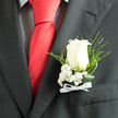 In cool, flattering light the ivory rose boutonnière presents a refined contrast to a smart charcoal jacket, the rose's velvety petals layered like soft snow against dark cloth. Nearby, the subtle texture of a checked shirt peeks under the lapel, and a crimson silk tie provides a bold vertical line that lifts the composition; together these tailored textiles set a sartorial stage for the flowers. Wisps of baby's breath cluster at the rose's base, their tiny white points like distant stars, imparting an air of innocence and quiet celebration. Fine strands of fern and twisted, glossy leaves arch around the bloom, creating rhythm and gentle movement that keeps the arrangement from feeling static. The white ribbon bow at the stem is narrow and neat, its smooth sheen echoing the tie's polish without competing for attention. Close inspection reveals the tactile chill of freshly cut petals and the faint, classic perfume of the rose-an intimate sensory note that complements the refined clothing textures. As a florist who knows the local rhythm from Tufnell Park Road to Junction Road, I consider how this boutonnière will read in photos taken outside the tube or along Dartmouth Park's quieter streets: composed, elegant, and confident. Flowers Tufnell Park crafts pieces like this to sit comfortably with both contemporary suits and more traditional morning coats, carrying a feeling of loyal devotion and ceremonial calm suitable for weddings, graduations, and other milestones where a single bloom can carry meaning.