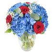 A luminous, garden-inspired bouquet sits in a simple clear glass vase, photographed against a clean white backdrop so each colour reads vividly. The arrangement is built from large, rounded clusters of sky-blue hydrangeas whose many delicate petals form a soft, cloud-like mass at the centre; the blue feels cool and watery, like a summer sky after rain. Nestled through these voluminous blooms are three deep red roses with velvety, tightly furled petals that offer a dramatic, passionate punctuation against the blue. Flecks of tiny white baby's breath drift between the larger flowers, bringing an airy, starry lightness that softens the composition, while muted, oval eucalyptus leaves and fresh green foliage thread through the bouquet, adding a subtle herbal scent and textural contrast. The vase has a gently rounded base and a slightly tapered neck that reveals the straight green stems submerged in clear water, a quiet detail that signals freshness. Bright, natural light-as one might see in a north-facing Tufnell Park kitchen or on a windowsill near Fortess Road-casts soft highlights across the petals, making the blues glow and the reds deepen. The overall mood is both celebratory and intimate: suitable as a centrepiece for a small anniversary supper, a patriotic display for a summer gathering, or a thoughtful gift to mark a birthday or milestone. As a florist's composition it balances volume, colour and scent so the arrangement reads beautifully from every angle and brings a feeling of bright, composed joy into a home in Tufnell Park or nearby Dartmouth Park.