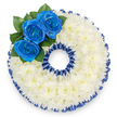 A bespoke funeral tribute formed unmistakably into the number '0', photographed from above on a clean white surface so every detail reads clearly. The body of the numeral is densely carpeted with hundreds of small white spray chrysanthemums, their tiny petals overlapping to create a cloud-like, tactile cushion that suggests calm and consolation. A narrow, pleated royal blue satin ribbon traces both the outer and inner rims of the zero, the ruffles catching a soft overhead light and producing a subtle scalloped silhouette that echoes the rounded shape. At the upper left curve a cluster of three deep royal blue roses provides the arrangement's emotional focal point: their tightly furled centers opening into velvety spirals, the blue hue almost luminous against the pristine white chrysanthemum field. Small, glossy dark green leaves sit tucked behind the roses, their cool tones adding depth and a whisper of natural freshness. The blue could be a deliberate tribute to a beloved uniform, a favourite colour, or a local club or society the recipient cherished. The chrysanthemum's gentle, earthy scent is almost imperceptible in the image but implied in the layered textures; the ribbon's sheen lends a formal, ceremonial finish. This custom number wreath is the kind of personal memorial we design for commemorations and wakes across Tufnell Park and neighbouring Dartmouth Park and Archway, created to mark an age or a special anniversary with clarity and quiet dignity. The overall mood is respectful and composed - a precise, thoughtful floral statement that balances softness with solemnity and reads as an intimate message of remembrance.