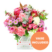 An abundant mixed bouquet of pink-toned flowers is artfully arranged in a clear, square glass vase and photographed against a pristine white background, giving the entire composition a fresh, modern feel. The arrangement is dominated by roses in shades of deep fuchsia, rich magenta, and gentle blush pink, some petals darkening elegantly at the tips, others unfolding in smooth pastel spirals. Interwoven among them are plush carnations in coral, peachy pink, and soft white, their finely ruffled petals providing a luxurious, almost fabric-like texture. Sprinkled throughout, petite daisy-style chrysanthemums in lavender and purple with bright green centres add a cheerful, slightly whimsical touch, as if echoing the vibrant, creative spirit found around Tufnell Park's cafés and side streets. Creamy white and softer-toned blooms help to balance the stronger pinks, ensuring the bouquet reads as harmonious rather than too bright. Lush greenery surrounds and supports the flowers: distinctive rounded eucalyptus leaves with a silvery cast, deeper green foliage, and a few subtle reddish-brown accents that bring warmth and depth, reminiscent of early evening light over nearby Gospel Oak. The bouquet sits neatly in a transparent vase with rounded corners, wrapped at its base by a decorative white band printed with repeating red hearts and the words all my love in a flowing red script, making the romantic message visible at a glance. In the lower right corner, a bold orange circular badge with white, capitalised text clearly states VASE INCLUDED, signalling that the arrangement arrives complete and ready to set down on a dining table, bedside, or office desk. The overall effect is joyful, tender, and perfectly suited for anniversaries, birthdays, Valentine's, Mother's Day, or simply sending heartfelt love across Tufnell Park and North London.