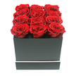 An opulent square box of red roses is the sole focus of this image, like a carefully considered gift waiting to be hand-delivered to a doorstep near Tufnell Park station. The arrangement consists of twelve perfectly preserved red roses, each one fully open and evenly sized, nestled side by side in three neat rows of four. Their petals are thick and velvety, coiling inward in intricate crimson spirals that capture subtle shifts of light from deep burgundy shadows to rich ruby highlights. The roses sit snugly within a matte black square box, their heads rising just above the rim so that the top surface appears densely carpeted with colour. The box itself has a contemporary, understated elegance: its smooth, untextured sides are broken only by a slim white band that wraps around the lower third, adding a refined accent reminiscent of minimalist interiors in North London apartments. The composition is set against a pure white background, with gentle, diffused lighting that softens any hard edges and lets the intensity of the red and black palette dominate the scene. Delicate shadows trace the contours of the petals and the crisp outline of the box, giving the arrangement a sense of depth without distraction. There are no ribbons, logos, or additional flowers, just the striking contrast of passionate red roses and sleek black packaging. The overall impression is one of sophistication and lasting sentiment, ideal for anniversaries, Valentine's celebrations, or heartfelt apologies sent across Tufnell Park, Kentish Town, or nearby Holloway, when words alone are not quite enough.