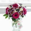 A vibrant mixed bouquet sits proudly in a clear, gently textured glass vase, placed against a softly glowing white background that feels like daylight pouring through a Tufnell Park window. The arrangement is dominated by large gerbera daisies in deep fuchsia and magenta tones, their velvety petals circling dark, almost chocolate-brown centres that pull the eye into the heart of the design. Around them cluster bicolour chrysanthemums, each petal tipped in rich purple that fades delicately into white, with bright yellow centres adding a cheerful spark. Creamy white spray roses, some fully open and others still in tight, hopeful buds, thread through the brighter tones, bringing a touch of classic romance and soft fragrance. Wisps of white gypsophila cloud between the larger blooms, creating a gentle, airy texture that feels like a breath of fresh air drifting across nearby Tufnell Park Fields. Deep green foliage peeks from beneath and between the flowers, grounding the composition and giving it a lush, garden-fresh feel, as if just gathered from a local London market. The low, jar-style vase is finished with a simple bow of rustic twine around its neck, from which hangs a neat white gift tag bearing a stylised pink flower logo and subtle lettering, hinting at a personalised message for a birthday, thank you, or just-because surprise. Soft natural light skims the glass and illuminates the petals, making the colours glow without harsh shadows, like a bouquet waiting on a bright kitchen windowsill off Fortess Road. The overall impression is one of joyful celebration and thoughtful gifting, the sort of fresh flower arrangement that could be delivered across Tufnell Park to lift someone's day and brighten a quiet corner of their home.