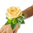 A delicate fresh wrist corsage rests gently on the inner wrist against a clean, white background, the kind of small, crafted elegance you might see carried from a Tufnell Park flat to a neighbourhood celebration. At its heart sits a single, fully opened rose in a warm peach-to-apricot hue; its soft, spiralling petals catch a subtle, diffused light and invite a gentle touch. Below and to the lower left of the rose are two bright lime-green button chrysanthemums - compact, almost spherical pom-poms whose tightly packed florets add playful texture and a lively contrast to the rose's velvety curves. The flowers are cradled on a bed of deep green foliage - broad, glossy leaves that read like salal or ruscus, giving the arrangement weight and a lush base. A distinctive variegated leaf, striped with pale and deep green, is artfully folded and curled so it sweeps from beneath the rose toward the right side of the wrist, lending a modern, architectural flourish that balances the soft florals. The corsage appears secured out of sight, likely with an invisible band or ribbon, so the composition reads as seamless and wearable. The overall palette of peach, lime, and rich greens feels quietly celebratory - suited to a wedding morning on Fortess Road or a prom evening after a walk through nearby Dartmouth Park - and carries a faint suggestion of rose fragrance and cut-green freshness. As a local florist's piece, it shows careful hand placement, fresh stems, and a refined restraint that lets each element speak: romantic, modern, and perfectly scaled to the wrist it adorns.