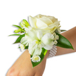A delicate wrist corsage unfolds like a quiet promise: a creamy ivory rose sits at the centre, its velvety petals unfurling with soft, luminous edges that catch the studio light. Nestled slightly below and to the left, a larger trumpet-shaped white bloom-evoking either an Alstroemeria or a small lily-opens to reveal a gentle green-yellow throat, lending a subtle, lively depth to the otherwise milk-white palette. Tiny, airy sprays of Baby's Breath drift through the composition like soft clouds, while several bright green buds and narrow, glossy leaves push through the blooms, suggesting the fresh possibility of new flowers to come. At the lower edge, a cluster of small white daisy-like florets with golden-yellow centres adds a playful beat to the arrangement's rhythm. The whole cluster rests against fair skin and is secured by a multi-strand band of lustrous white pearls; the elastic bracelet wraps smoothly, the pearls' cool sheen mirroring the pale blossoms and adding a tactile note of refinement. The background is clean and white, allowing the nuanced greens and creams to read clearly, and a faint, powdery floral scent seems almost present in the air. As a piece meant for moments of ceremony-proms, weddings, or a quiet vow exchanged beneath the trees of Dartmouth Park-this corsage balances timeless elegance with a whisper of youthful joy. A Tufnell Park florist's touch is evident in the careful spacing and the way each leaf cradles a bloom, making this wristlet feel at once handcrafted and composed for celebration.