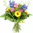A joyful mixed bouquet is pictured upright against a pure white background, its colours and textures standing out as clearly as if displayed in the window of a Tufnell Park florist. In the centre, open pink roses show layers of gently curving petals, giving a soft, romantic heart to the design. Clustering around them, bright yellow gerbera daisies with dark brown centres form bold, circular bursts of sunshine, adding a lively, upbeat note that recalls the friendly atmosphere around Tufnell Park station. Tall stems of vivid blue delphiniums rise behind and above the rounder blooms, their many small flowers lined along each stem to create graceful vertical lines and a cool, almost sky-like contrast to the warmer tones below. The bouquet is generously cushioned with greenery: feathery fern fronds peek from the edges, while shiny, deep green leaves offer a sturdy base of colour, making the pinks, yellows, and blues appear even more intense. Tucked throughout are bright green, textured filler flowers and tiny buds, contributing volume and an intricate, garden-fresh feel. At the lower part of the bouquet, broad tropical-style leaves, some with faint pale green striping, are carefully curved and wrapped to form an elegant leafy collar, giving the arrangement a neat, modern silhouette. Beneath this, the exposed stems are drawn together and tied with a soft, pale wrap, hinting at a hand-crafted, hand-tied finish. The overall impression is of a fresh, celebratory bouquet ready for same-day delivery across Tufnell Park, perfect for birthdays, congratulations, or simply brightening a flat near the quiet streets by Dartmouth Park Hill.